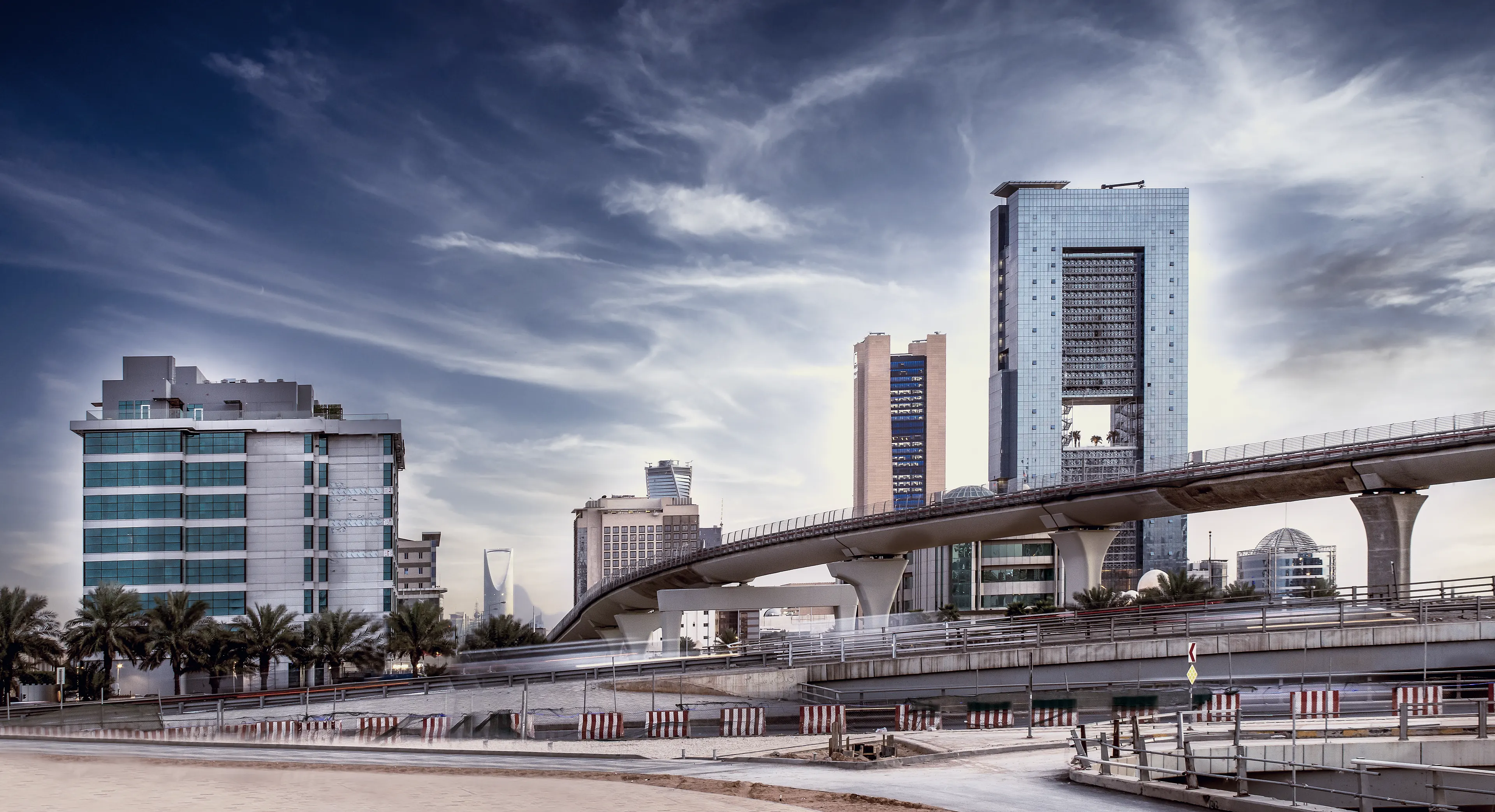 Riyadh by day: the towers of the central business district under a dramatic sky, a highway bridge curving past.